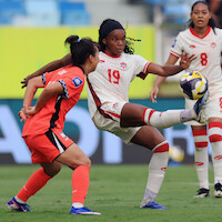 Soccer match action shot with a player in a white and red uniform kicking a ball while a player in an orange uniform attempts to block; outdoor stadium, other players and audience blurred in background.