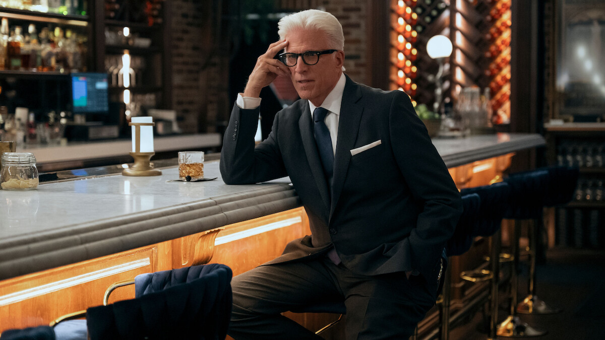 Older man in a dark suit sits at an elegant bar with modern lighting and upscale decor, looking thoughtful and composed.
