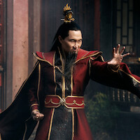 A man in ornate red and gold robes with a flame-shaped crown stands in a traditional Asian architectural setting, striking a dynamic pose with an intense expression.