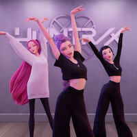 Three animated women in a modern dance studio pose gracefully with raised arms, wearing stylish black and pink outfits, wooden floor, and a wall with the "HUNTR/X" sign in the background.