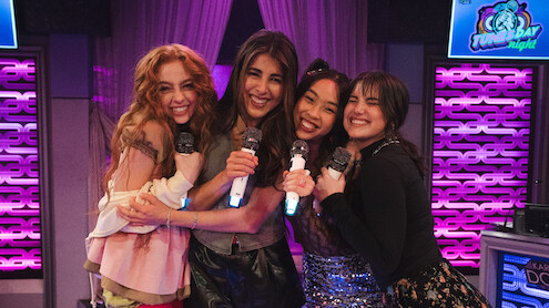 Four young women smiling and hugging while holding microphones on a brightly lit karaoke stage with purple lighting and decorative neon panels, enjoying a fun night out together.