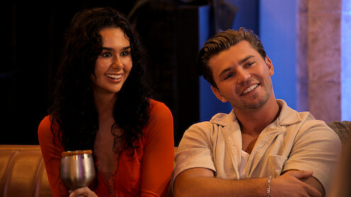 Two young adults sitting on a couch indoors, smiling and looking toward someone off camera. The woman holds a drink, both appear relaxed. Warm lighting suggests a social, casual atmosphere.