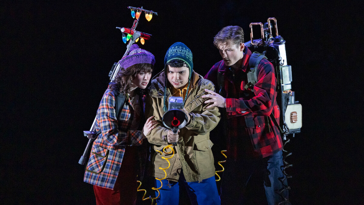 Three young people in winter clothes, holding ghost-busting equipment with glowing lights, stand closely together in a dark, dramatic setting, appearing focused and ready for action.