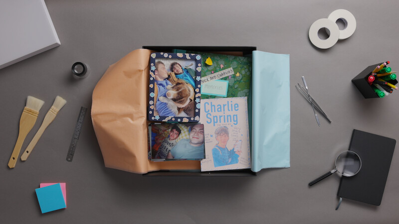 Scrapbook gift box with photos, book titled "Charlie Spring," and keepsakes on a desk. Surrounding items include paintbrushes, ruler, tape, pens, sticky notes, magnifying glass, scissors, and colored paper. Creative workspace setting.