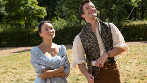 A woman and man in period costumes stand outside on a sunny day, smiling and looking up while the man flies a kite.