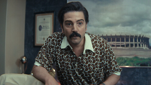 Man with dark hair and mustache in patterned shirt leans forward in an office with framed certificate on wall and large stadium photo in the background.