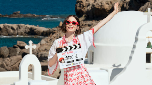 Woman wearing red sunglasses and a patterned dress stands smiling with one arm raised, holding a film clapperboard, in front of white buildings with domes and crosses by a rocky seaside under a blue sky.