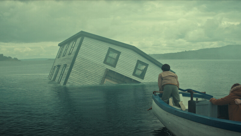 Two people in a small boat approach a partially submerged house tilted in the middle of a misty lake under a cloudy sky, with forested hills in the background.