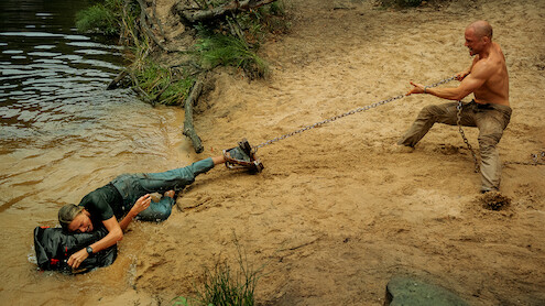 A shirtless man on sandy shore pulls a chain attached to a person partially in water, who is holding a black bag, with trees and water in the natural outdoor background.