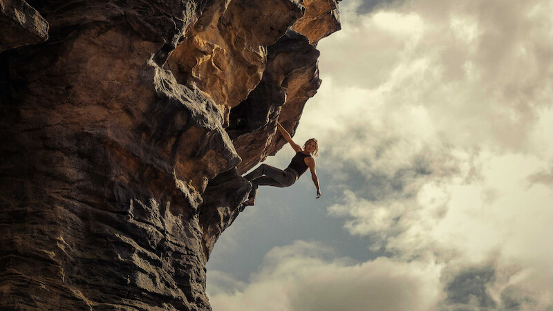 A rock climber scaling a steep, overhanging cliff outdoors under a cloudy sky, demonstrating strength and balance in a dramatic natural setting.