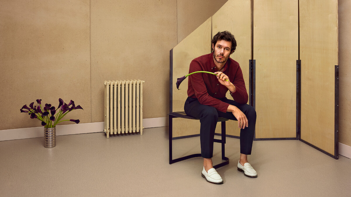 Adam Brody está sentado em um banco segurando uma flor copo-de-leite preta