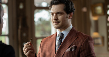 In a still image from the series 'The Tailor,' a man in a burnt sienna pinstripe suit jacket and a patterned brown tie holds a needle.