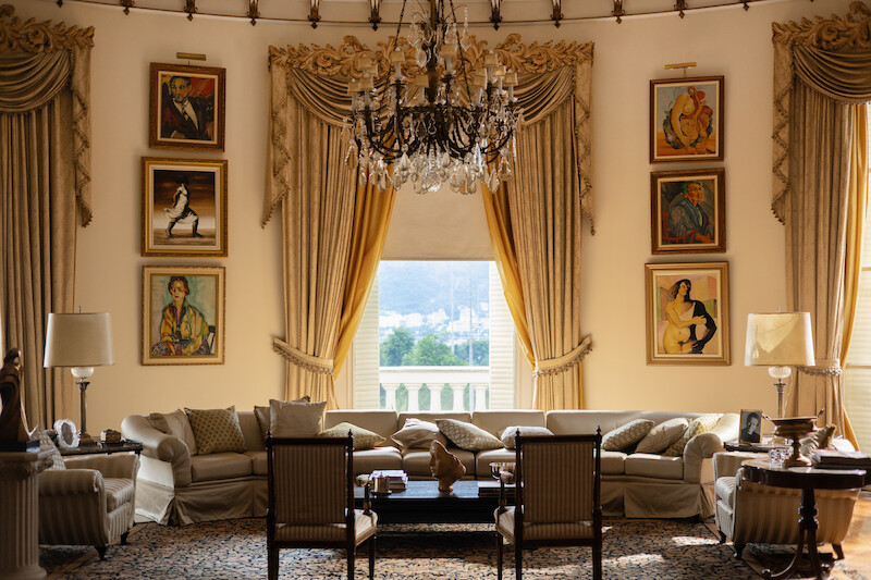 Uma grande sala oval tem um sofá centralizado com uma mesa de centro à sua frente. Ao fundo, cortinas douradas adornam as janelas e quadros empilhados um sobre o outro decoram as paredes. No teto, centralizado, há um grande lustre antigo