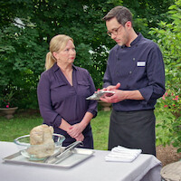 Two people in chef attire stand outside by a table set with cooking ingredients and utensils, discussing a recipe in a lush garden setting.