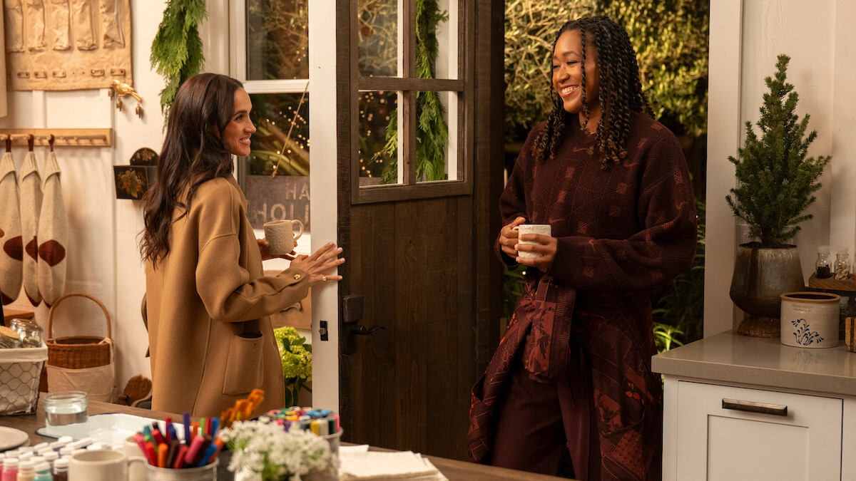 Two women smiling and talking in a cozy kitchen decorated for the holidays, holding mugs. Warm lighting, craft supplies on the counter, and greenery create a welcoming, festive atmosphere.