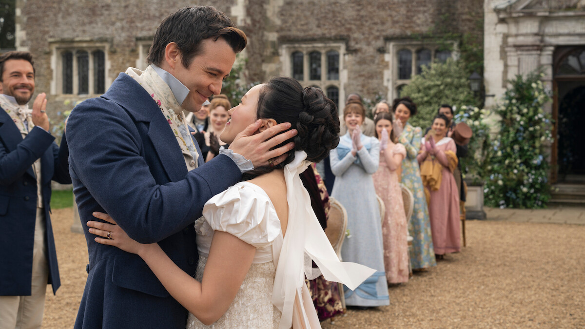 A bride and groom in period costumes embrace and smile outdoors, surrounded by cheering guests in Regency-era attire, with an old stone building and greenery in the background.