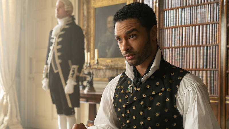 A man in Regency-era clothing sits in a richly decorated library with bookshelves and paintings, while a servant stands in the background near a window and candle holder.