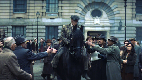 Man in vintage clothing rides a black horse through a crowd in front of an old stone building; the atmosphere is tense and dramatic, with people reaching out and reacting emotionally to his presence.
