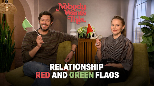 Adam Brody (Noah) and Kristen Bell (Joanne) sit on a couch in a cozy living room, smiling and holding green and red flags, with text about "Relationship Red and Green Flags," creating a lighthearted and conversational mood.