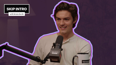 Louis Partridge smiles while speaking into a microphone during an interview on "Skip Intro with Krista Smith."