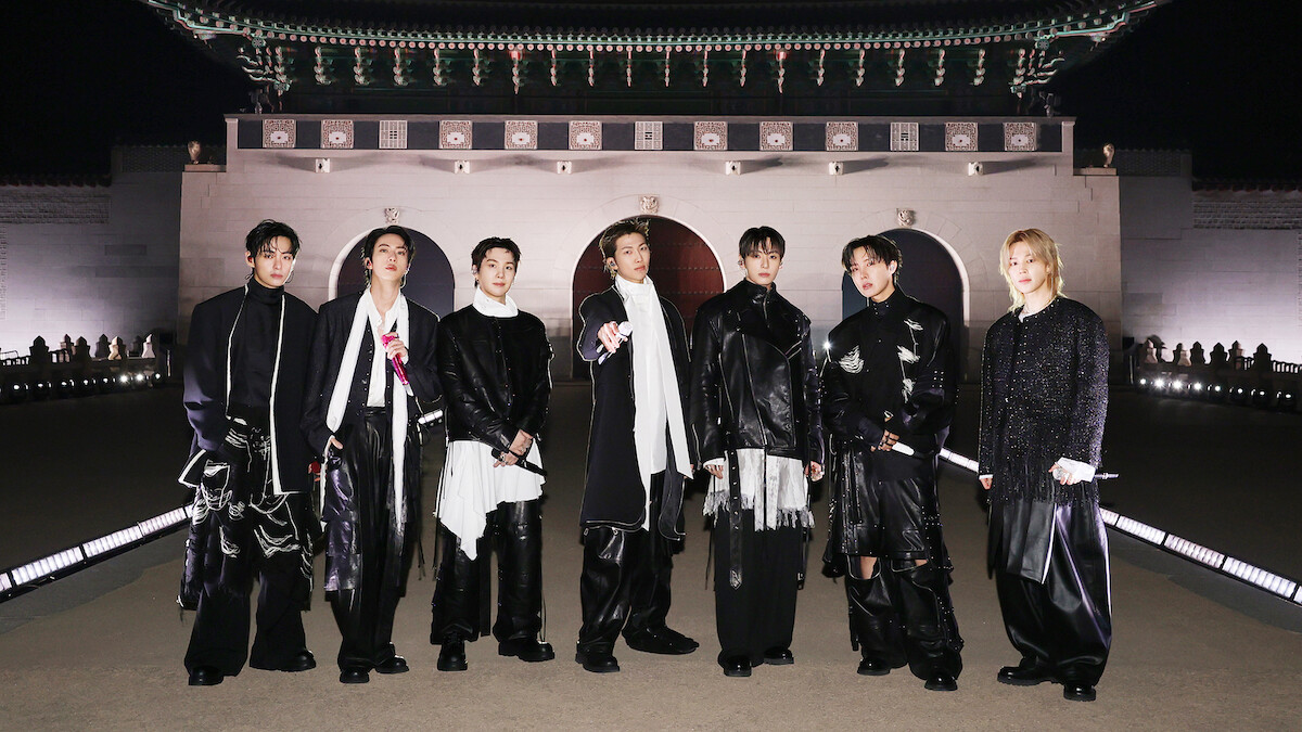 V, JIN, SUGA, RM, JUNG KOOK, J-HOPE, and JIMIN dressed in fashionable black and white outfits stand outdoors at night in front of an ornate, traditional East Asian building with arches and a large decorative roof.