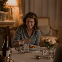 Woman smiling at a dinner table in a softly lit, elegant room with flowers, wine, and food, creating a warm and intimate dining atmosphere.