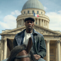 Man in a cap riding a horse in front of a historic domed building with a French flag, under a blue sky. The setting is urban, and the architectural style is classical.