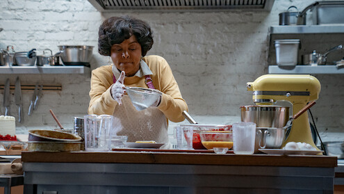 A woman in a kitchen wearing an apron and gloves, concentrating as she prepares food. The kitchen has a stand mixer, various bowls, utensils, and ingredients on a wooden countertop, with shelves and cookware in the background.