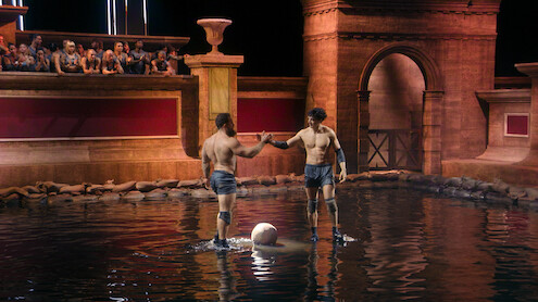 Two men in athletic gear stand in a shallow arena pool, shaking hands next to a ball, with spectators watching from seats above in a setting resembling ancient Roman architecture.
