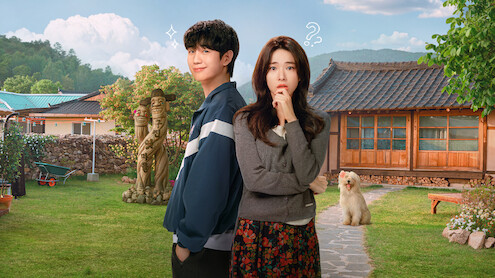 A young man and woman stand in a sunny, rural yard with traditional wooden house, lawn, garden statues, and a fluffy dog, mountains in the background, blue sky with clouds, both subjects displaying thoughtful and playful expressions.