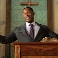 Man in a suit stands at a wooden pulpit with open arms, smiling in a church with a Bible and cross in the background, green and beige walls, and natural lighting.