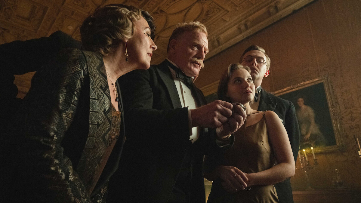 Four well-dressed people in formal evening attire stand close together in an ornate, dimly-lit room with a decorated ceiling and classical artwork, appearing to discuss something intently.