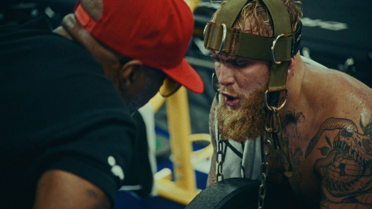 Jake Paul uses neck exercise equipment under the watchful eye of his trainer.