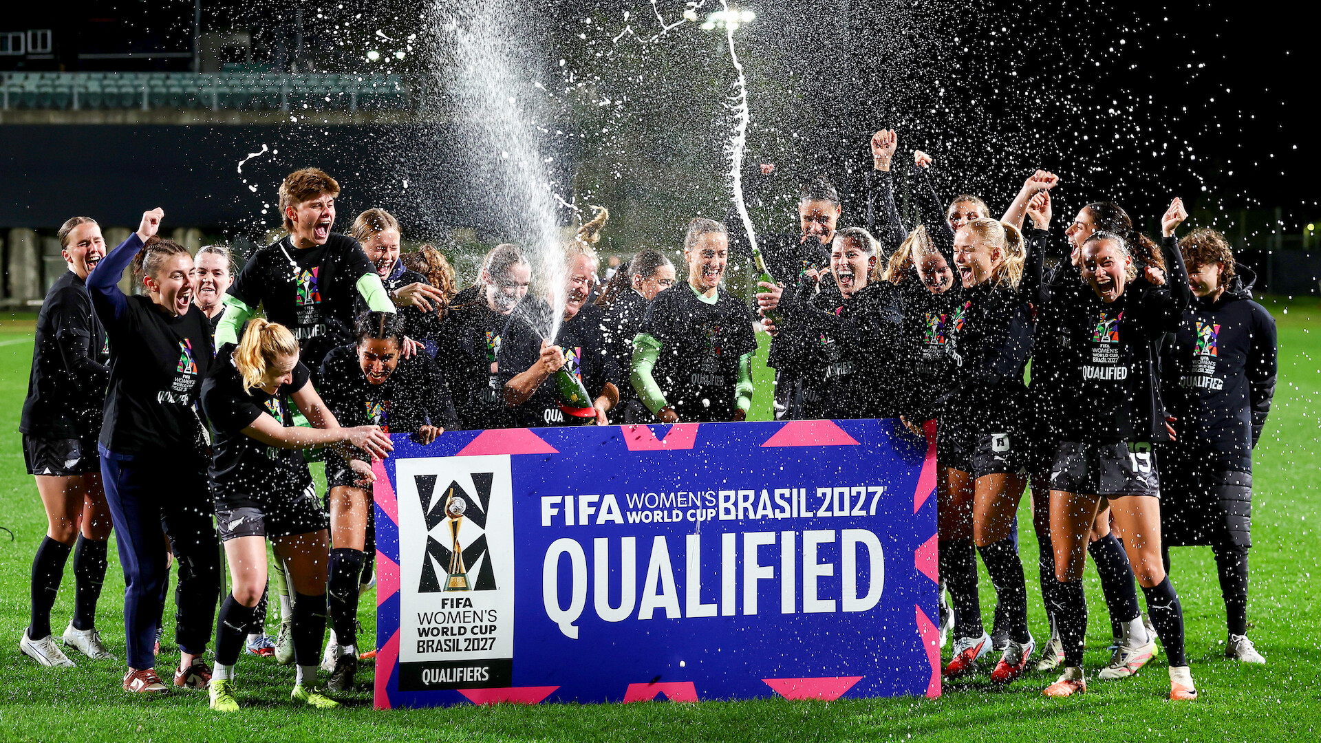 Women's soccer team celebrates at night on a field, spraying champagne and cheering behind a large sign reading "FIFA Women's World Cup Brasil 2027 Qualified." Bright stadium lights and festive atmosphere.