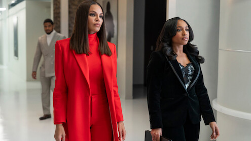 Crystle Stewart as Mallory and Taylor Polidore Williams as Kimmie in business attire, one in a red suit and the other in a black suit, walk confidently down a modern office hallway, with a man in a grey suit walking behind them.