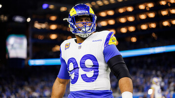 Football player in Los Angeles Rams uniform and helmet with number 99 stands on the field in a brightly lit stadium during a game, with a crowd visible in the background.