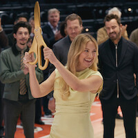 Woman in yellow dress holding large ceremonial scissors, preparing to cut a ribbon at an indoor event with smiling people clapping in the background, suggesting a celebration or grand opening on a basketball court.