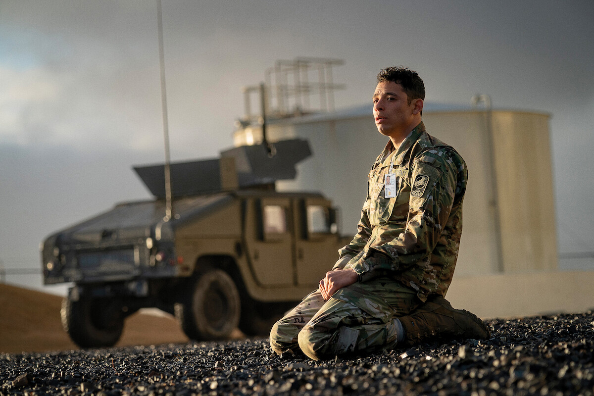 Um soldado de uniforme camuflado se ajoelha em um terreno rochoso perto de um veículo militar. Ao fundo, um tanque industrial aparece sob o céu nublado, em uma imagem de clima contemplativo e solene.