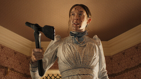 Woman in an old-fashioned white dress, splattered with blood, holding an axe inside a Victorian-style room with patterned wallpaper and a high ceiling, looking intense and determined.