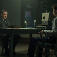 Two men sit across a table in a dimly lit interrogation room, one wearing a suit and the other connected to a polygraph machine, microphones and recording equipment between them.
