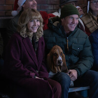 Smiling woman and man sit closely on a bench with a basset hound dog, dressed warmly in winter coats, indoors against a brick wall, with festive holiday atmosphere and people in background.