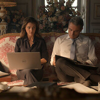 A woman with a laptop and a man reviewing documents sit on an ornate sofa in an elegant room decorated with flowers and classical furnishings, engaged in serious work.