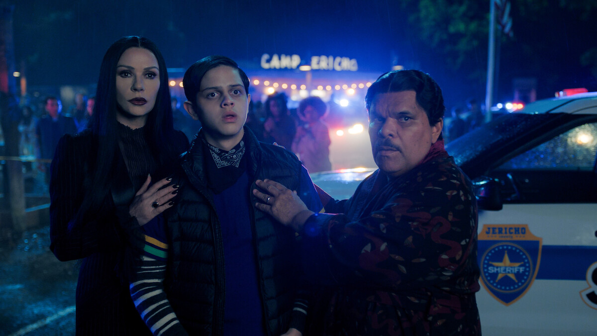 Catherine Zeta-Jones as Morticia Addams, Isaac Ordonez as Pugsley Addams, and Luis Guzmán as Gomez Addams stand in front of a police car in 'Wednesday' Season 2.