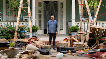 A man stands on a porch surrounded by construction materials, debris, and wooden scaffolding in front of a house. The setting suggests renovation work in progress, with scattered tools and building supplies.