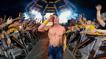 Shirtless wrestler saluting and walking down a ramp, surrounded by cheering fans at a brightly lit arena event, with colorful lights and stage setup in the background.