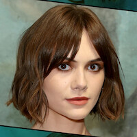 A person with short brown hair and bangs, neutral expression, photographed in a studio setting with a muted, artistic background.