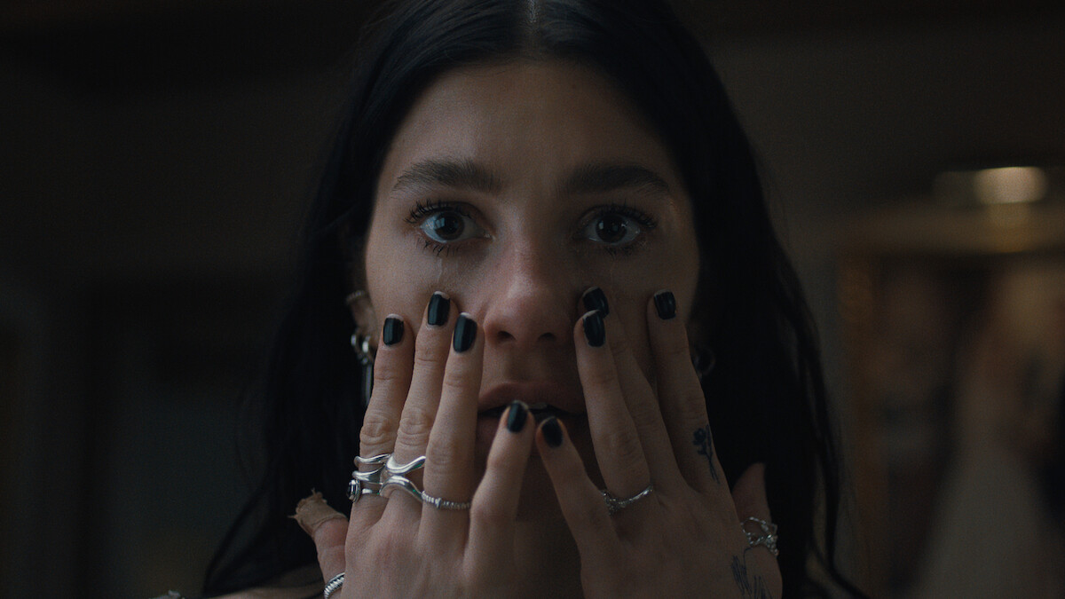 A woman with dark hair and black nail polish holds her hands to her face indoors, wearing multiple rings and looking surprised or concerned in a dimly lit setting.