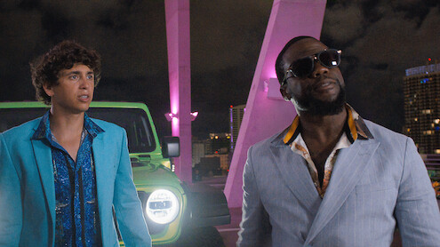 Two men in stylish suits stand in front of a bright green car on a city rooftop at night, with neon pink lights and skyscrapers in the background.