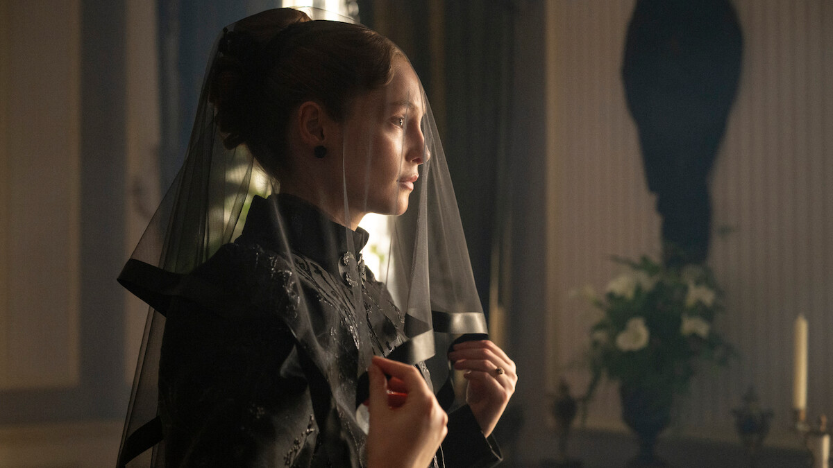 Woman in black mourning attire with a veil, standing indoors in a dimly lit, elegant room with flowers and candles in the background, suggesting a somber or formal occasion.