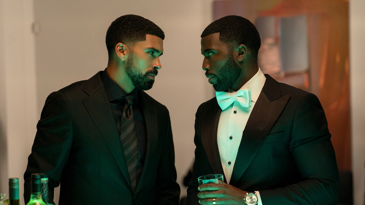 Two men in formal black suits stand close, engaged in a serious conversation at an indoor event. One holds a drink, bottles are visible on the table, and ambient green lighting enhances the elegant, upscale setting.